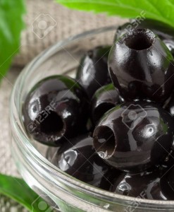 Detailed macro view of the black olives, pitted marinated in a glass bowl on a background of green leaves with shallow depth of field